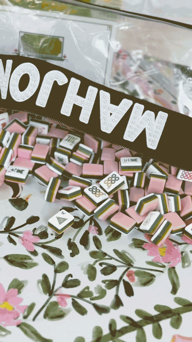 A package of Mahjohng game pieces on a floral tablecloth, with someone pouring rose.