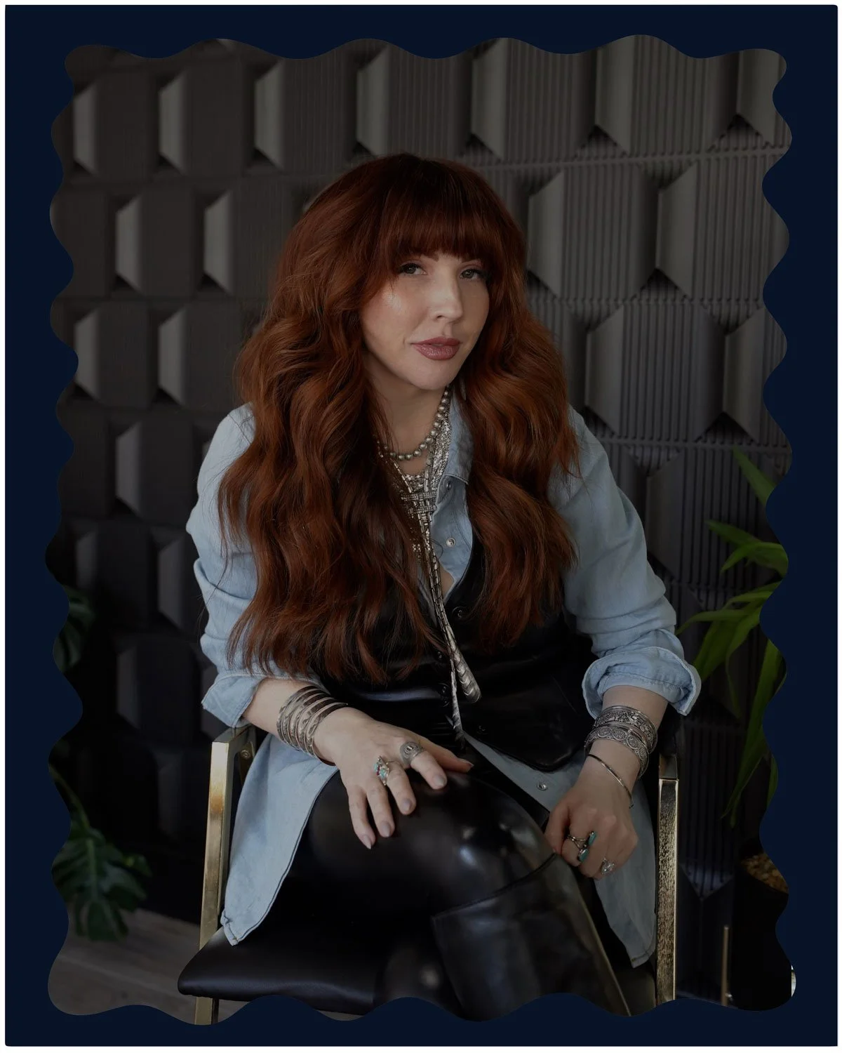 A woman with long, wavy red hair and bangs sitting in front of a gray, patterned soundproof wall, wearing layered jewelry, a denim shirt, a black top, and black leather pants.