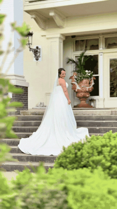 A bride in a white wedding gown standing on outdoor steps of a house, with a large flower urn and windows in the background.