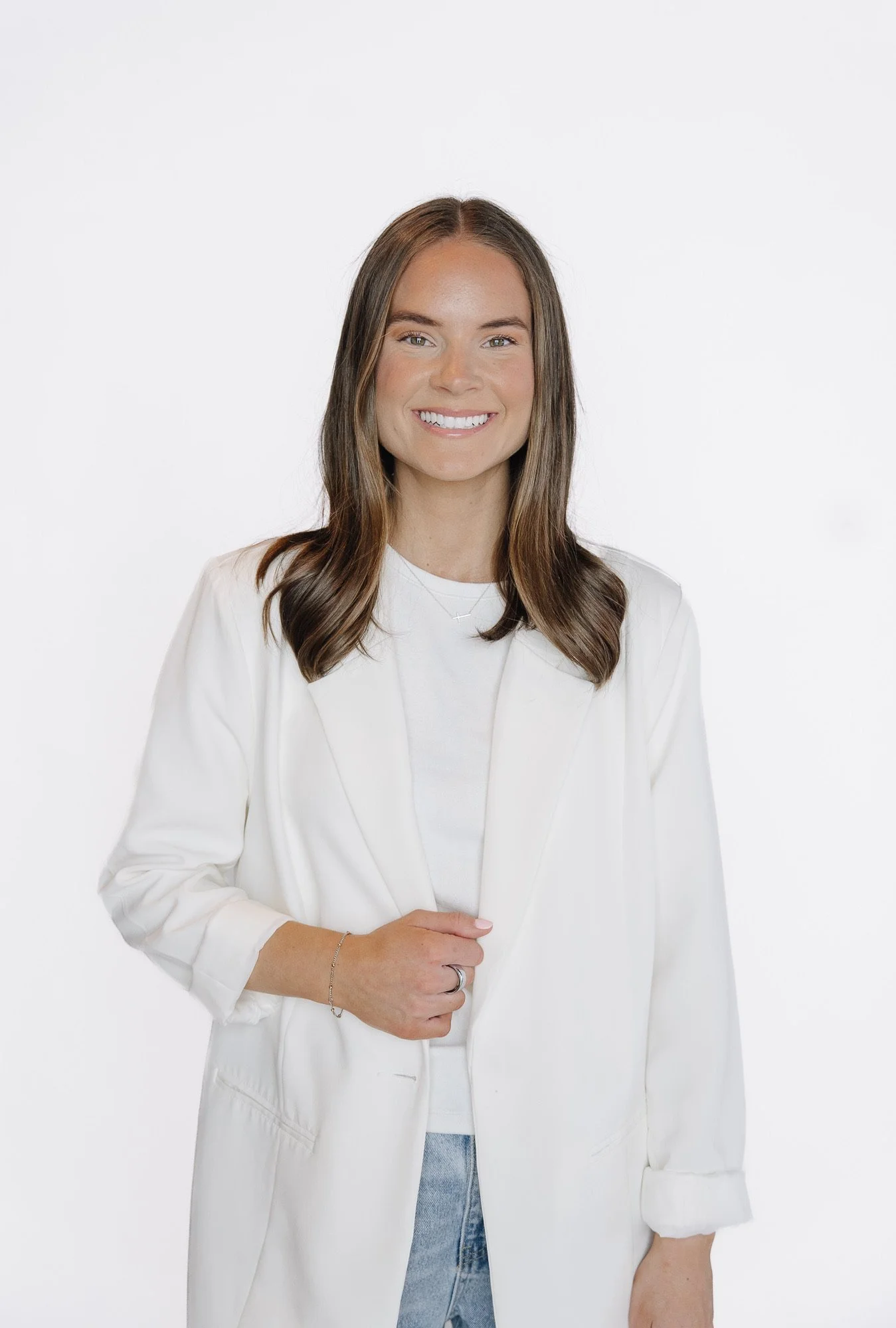 Smiling woman with long brown hair, wearing a white blazer over a white top, standing against a plain white background.