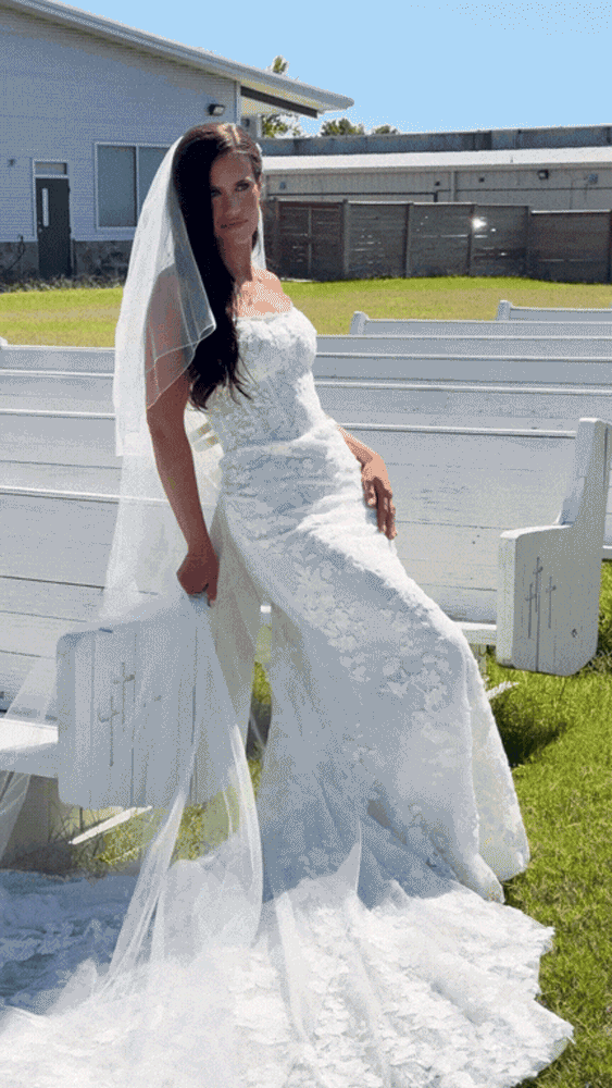 A woman in a white wedding dress and veil posing outdoors on a sunny day.