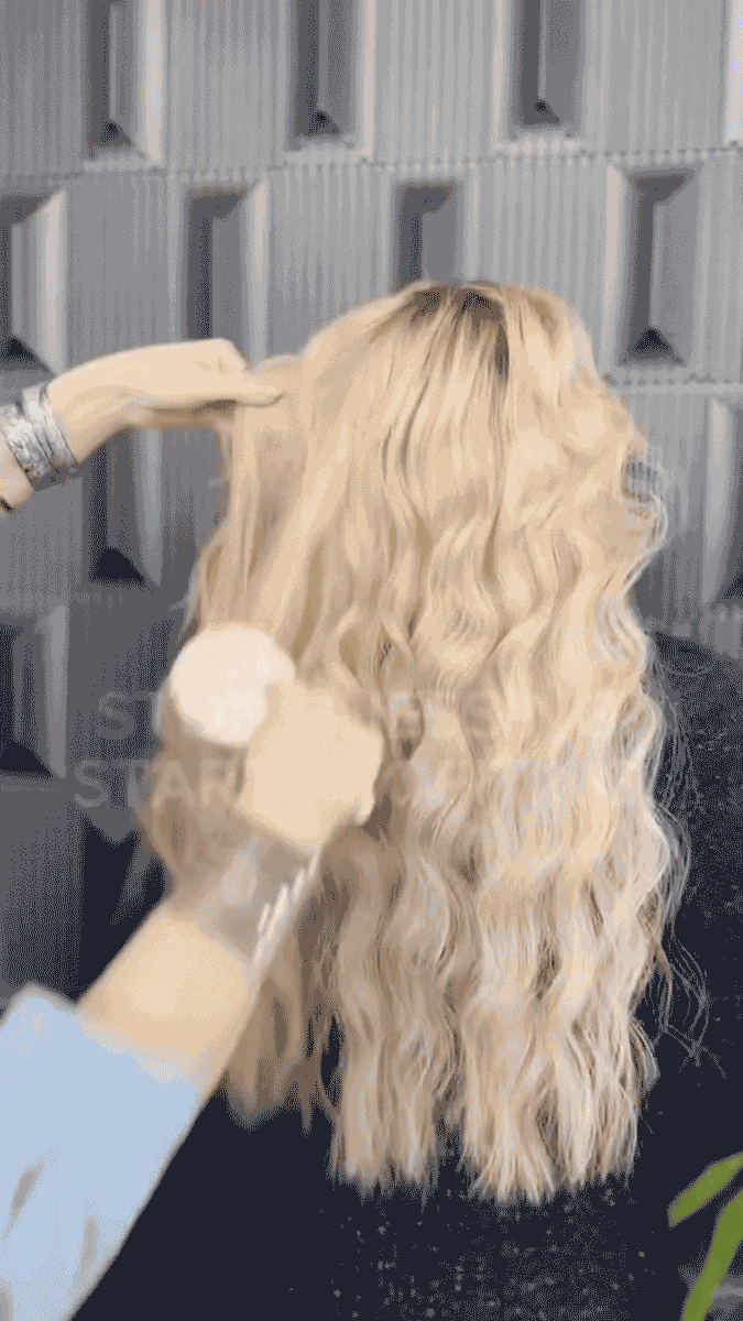 A person styling long, wavy blonde hair on a mannequin head in a salon or studio setting.