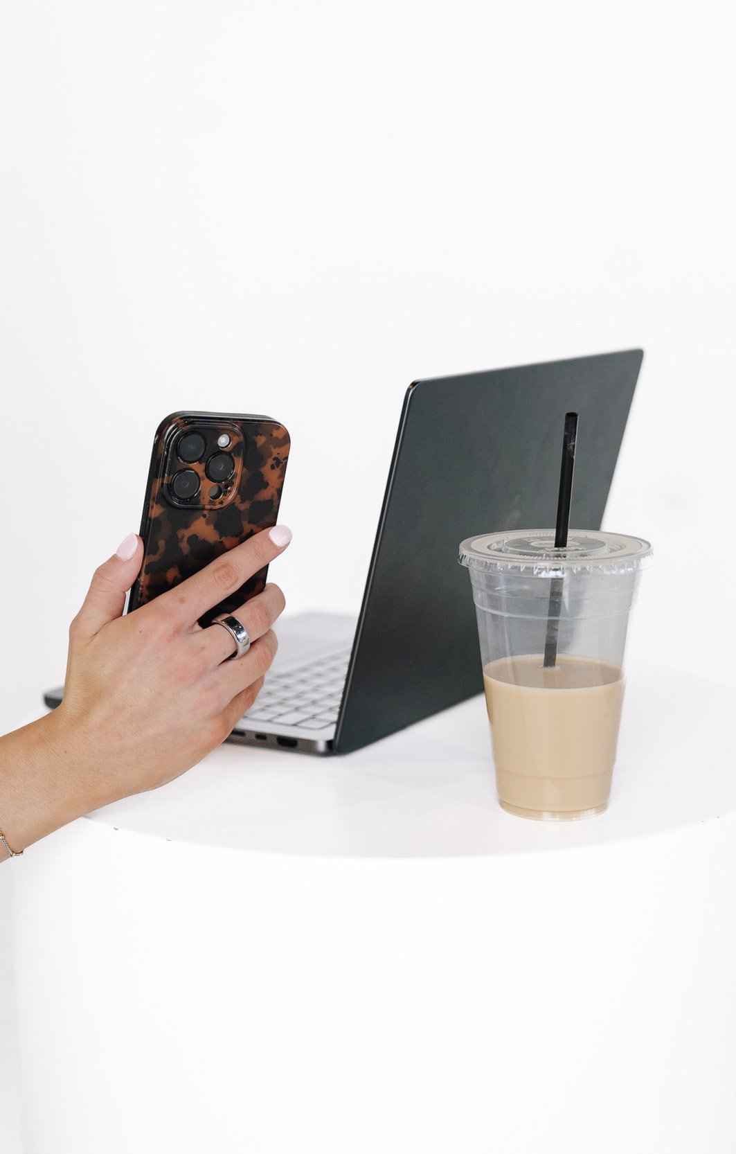 Hand holding a smartphone with a tortoiseshell case, a silver ring on the ring finger, next to a laptop and a clear plastic cup with iced coffee and a black straw on a white surface.