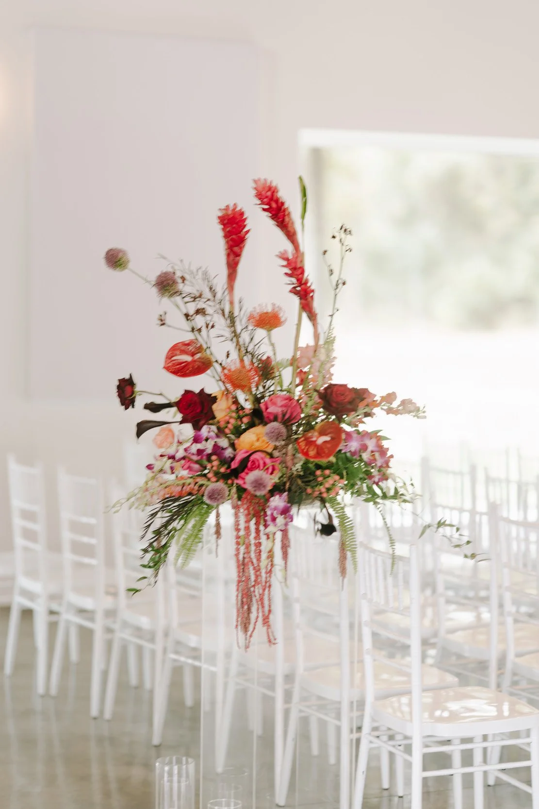 A tall flower arrangement with pink, red, and orange flowers on a stand in a bright room with white chairs and large window.