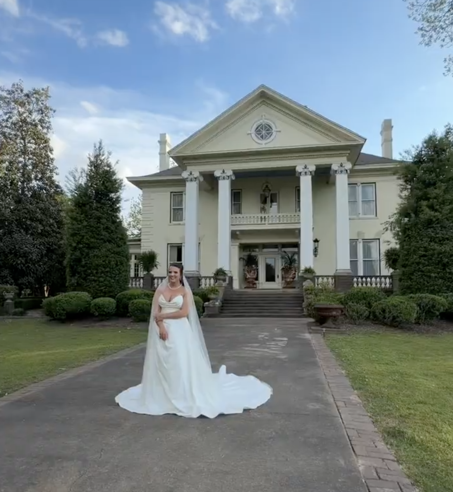 Bride in a white wedding dress standing on a driveway in front of a large, white, two-story house with columns and a balcony, surrounded by green trees and a grassy yard.
