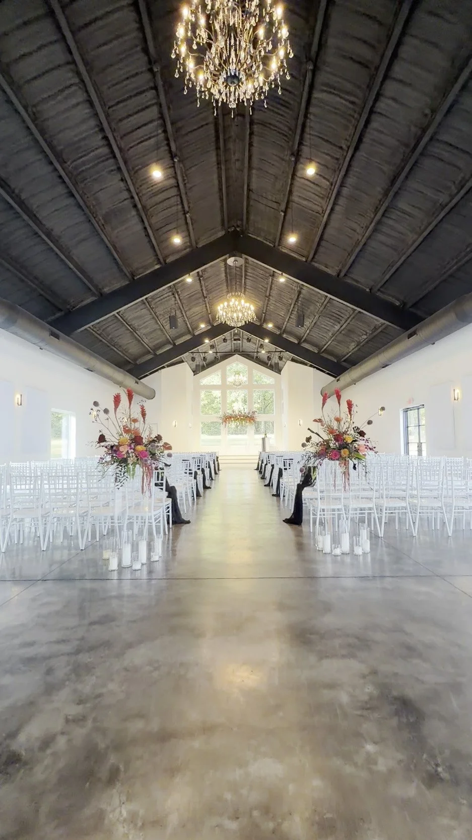 Indoor wedding venue with long aisle, white chairs, floral decorations, and chandeliers hanging from a dark wooden vaulted ceiling.