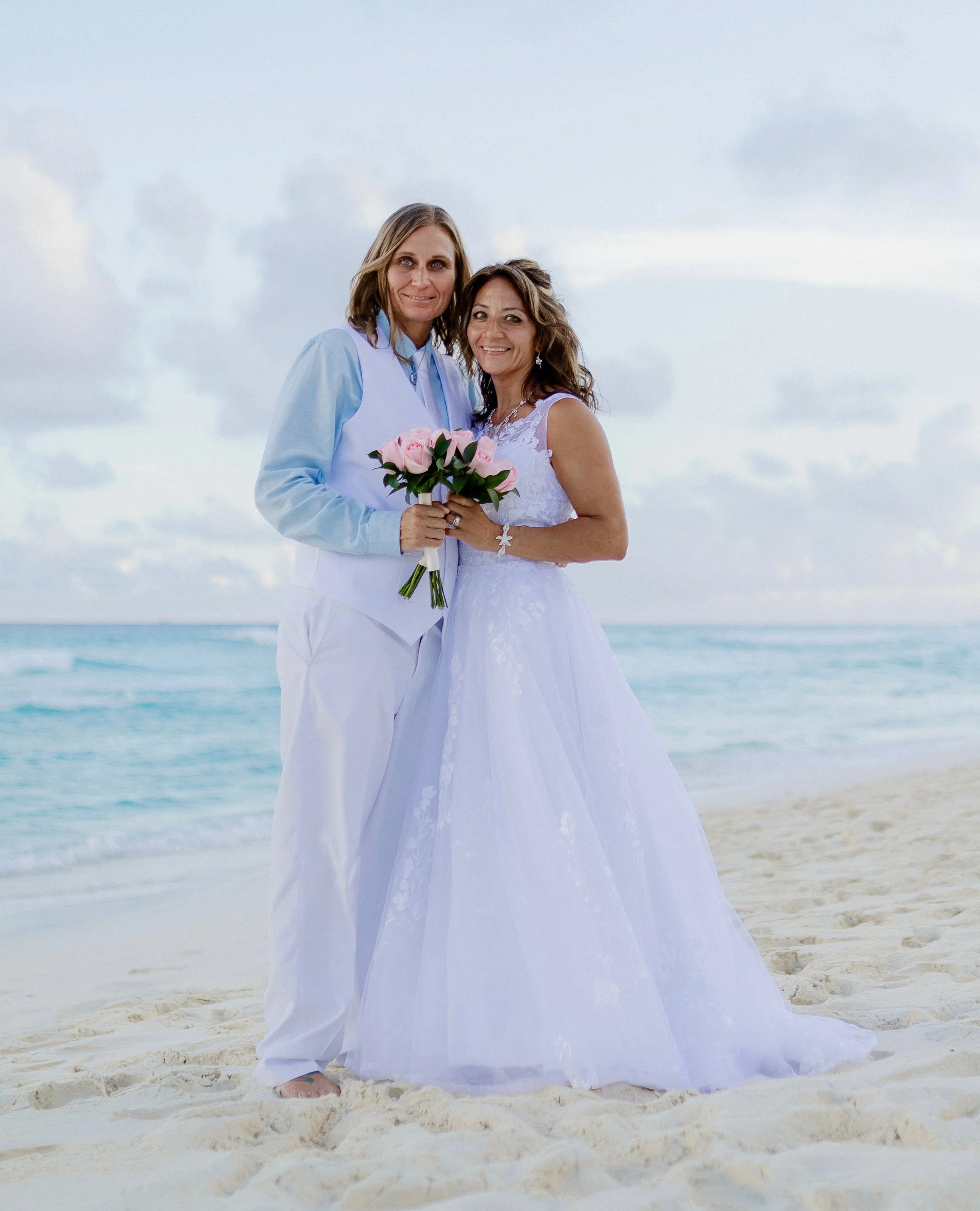 Two women in wedding attire holding pink roses during their beach elopement ceremony.