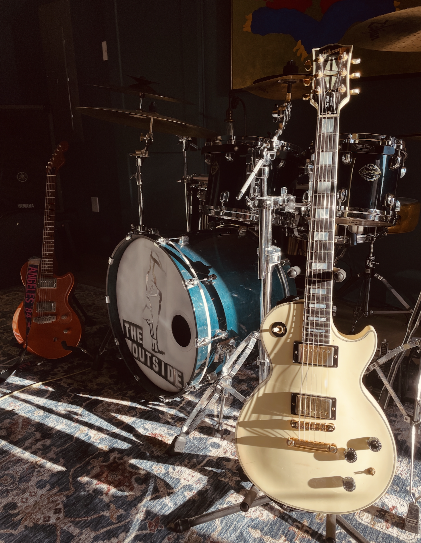A drum set with black drums and cymbals, an electric guitar leaning against the drums, a smaller orange guitar nearby, and sunlight casting shadows on a patterned rug.
