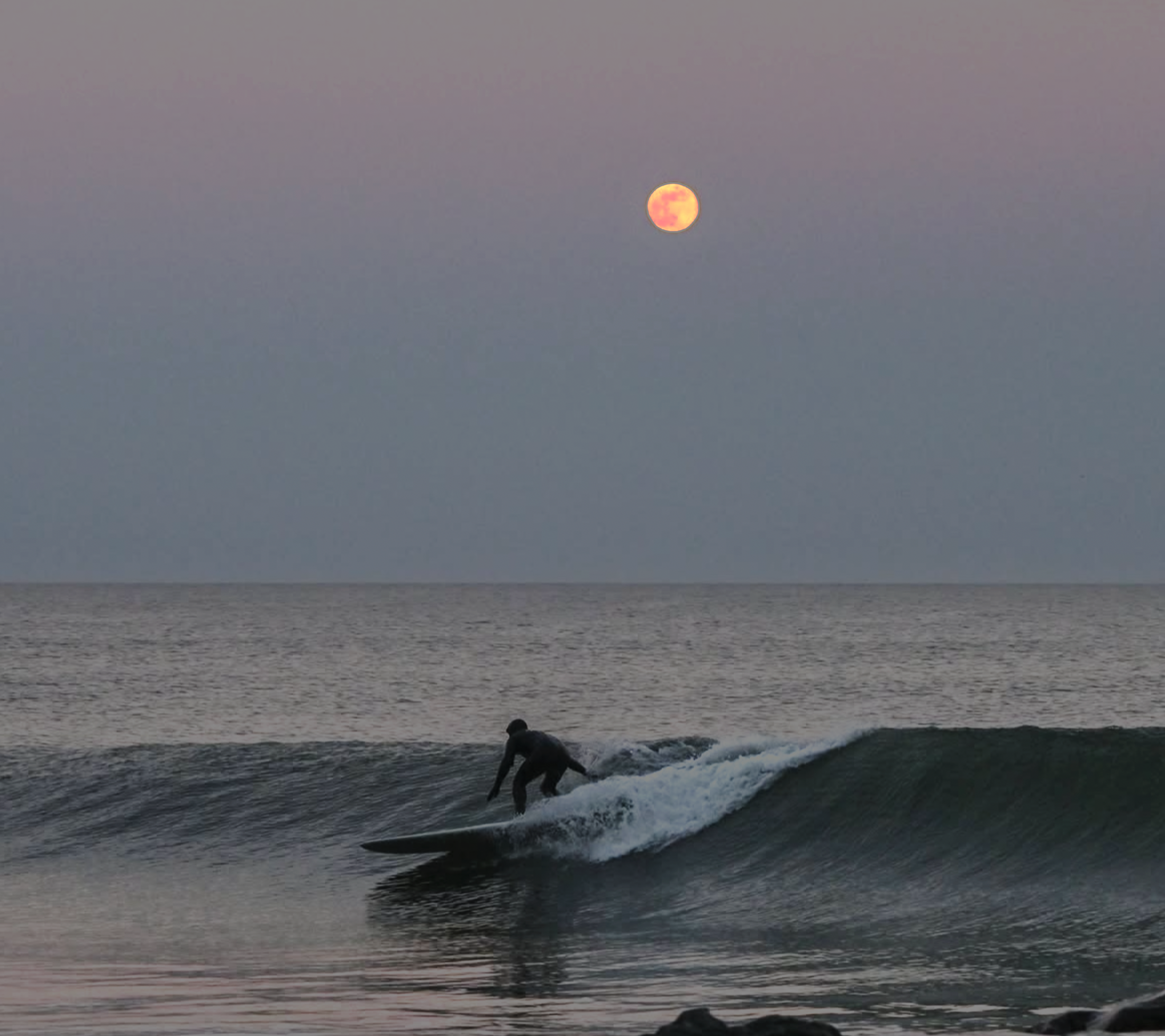 A person surfing on a wave in the ocean at dusk with a large, orange full moon in the sky.