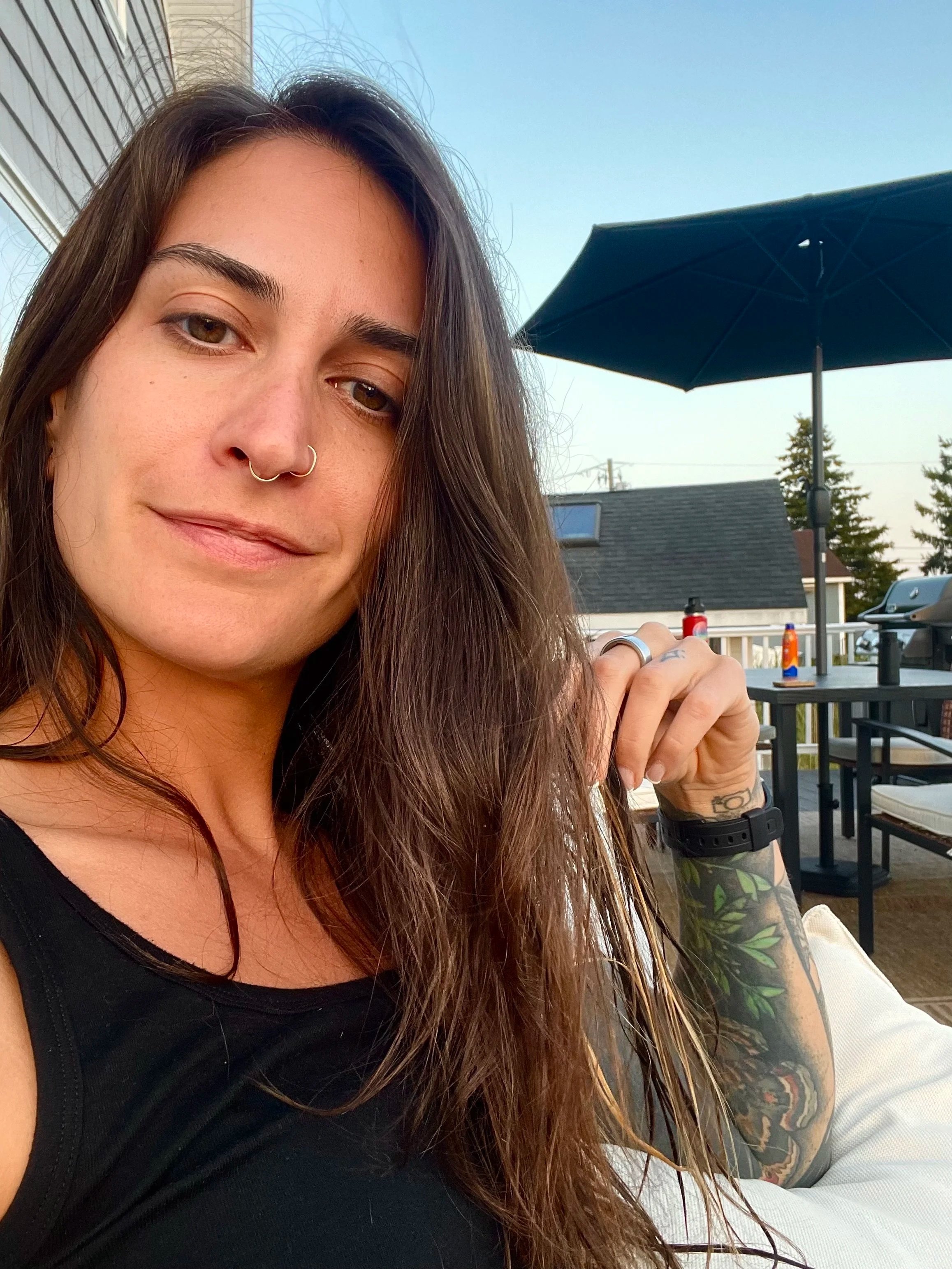 Close-up of a woman with long brown hair, styled with a middle part, wearing a septum ring, a black top, and a tattooed arm. She is outdoors on a patio with a black umbrella, some tables, and a house with a roof and trees in the background.
