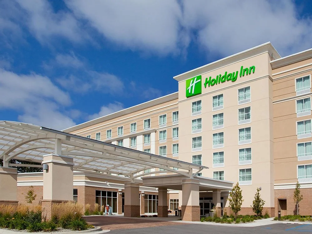 Exterior view of a Holiday Inn hotel with a covered entrance, trees, and a partly cloudy blue sky.
