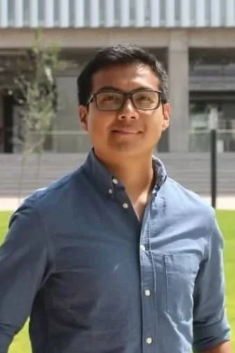 A young man with glasses wearing a blue button-up shirt, standing outdoors with modern buildings and greenery in the background.