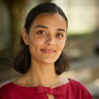 Young woman with dark hair wearing a red blouse, outdoors with greenery in the background.