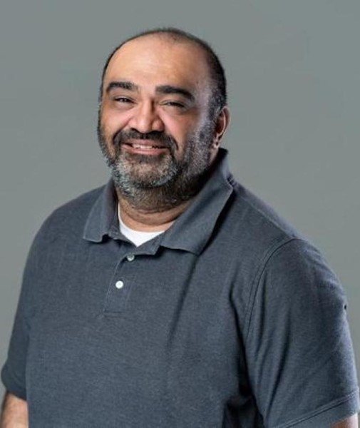 A middle-aged man with a beard and short hair, wearing a dark gray polo shirt, smiling at the camera against a plain gray background.