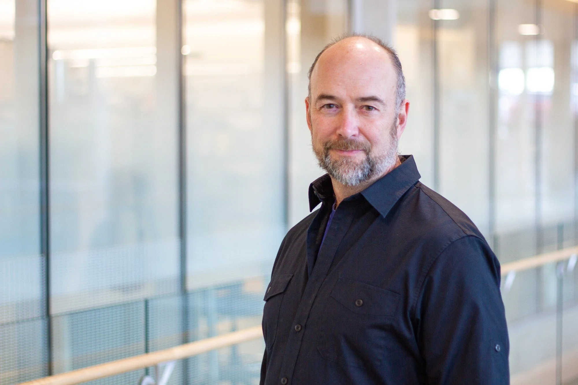 A middle-aged man with a beard and receding hairline posing indoors with a glass wall in the background.