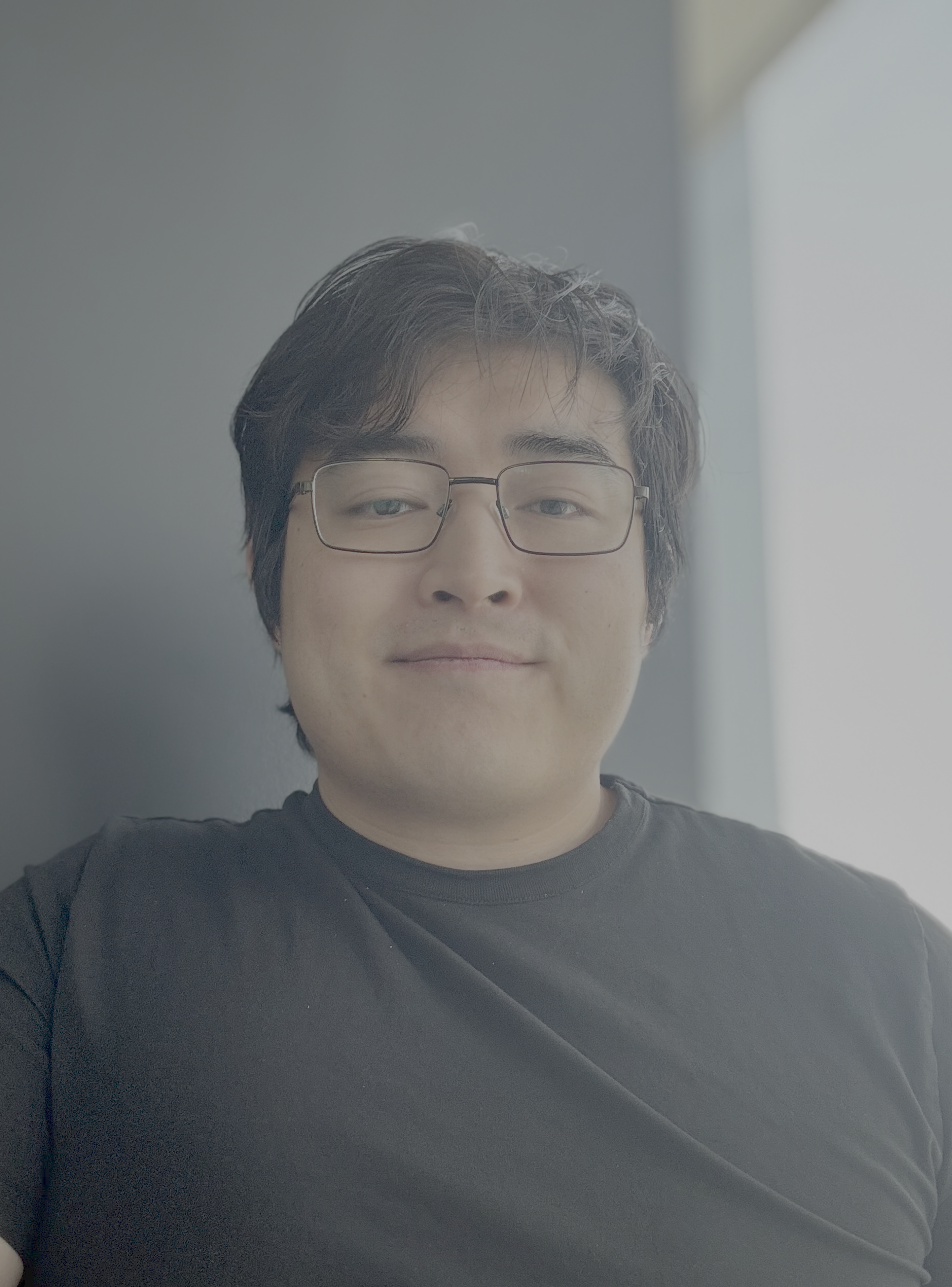 A young man with glasses and short, dark hair smiling softly in front of a window.