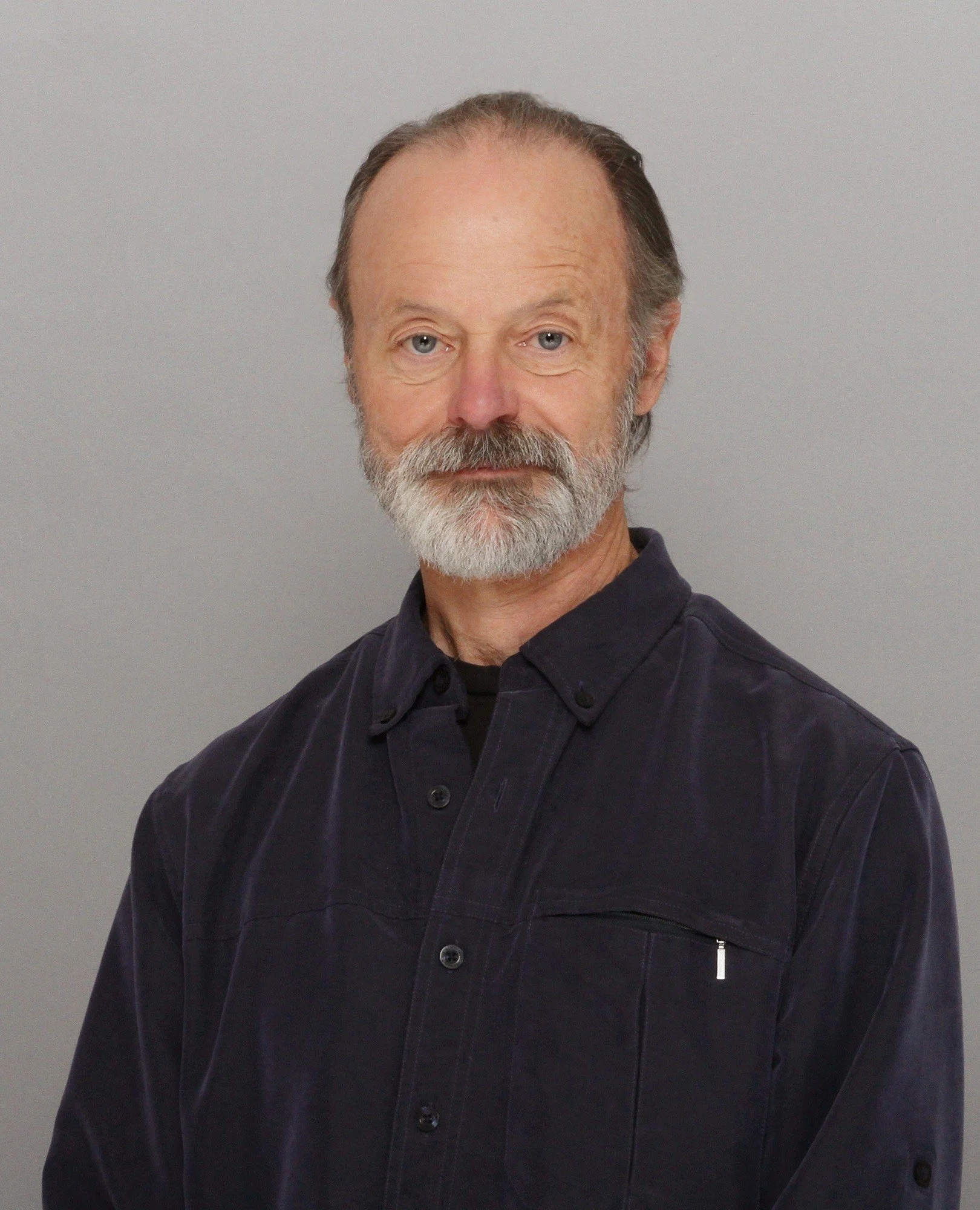 A middle-aged man with a beard and mustache, wearing a black button-up shirt, posing against a plain light gray background.