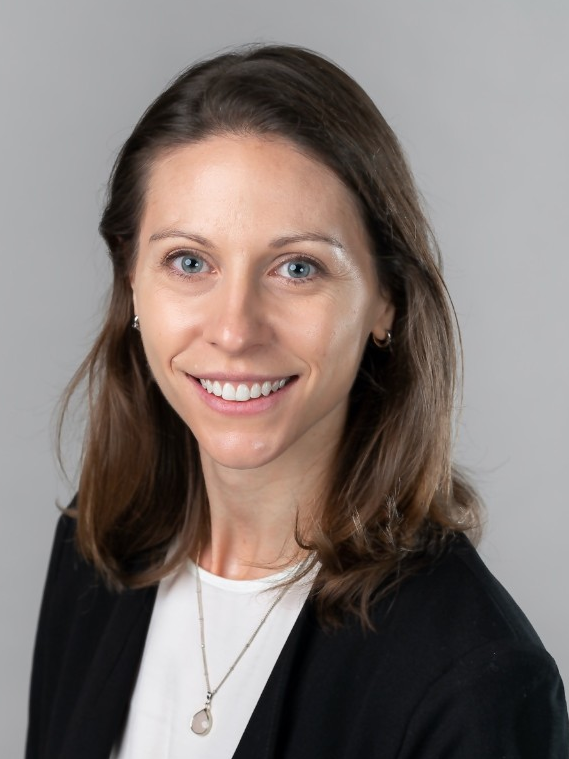 A woman with light skin, shoulder-length brown hair, and blue eyes smiling at the camera, wearing a black blazer over a white top and a necklace, against a plain gray background.