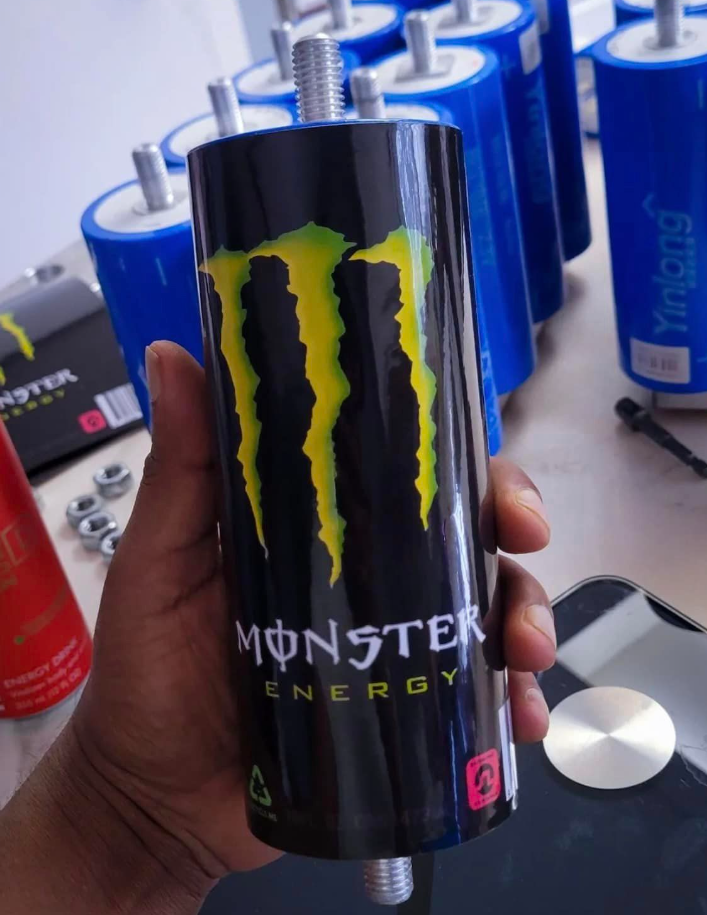 Person holding a black can of Monster Energy drink with bright green and yellow logo. Several blue cans are visible in the background.
