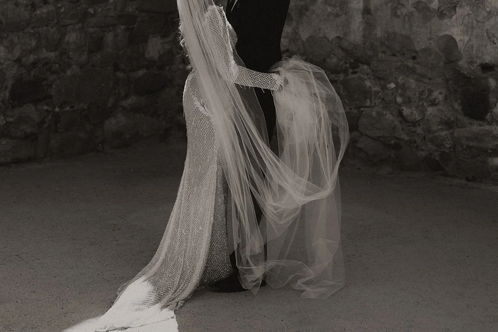 Close-up of a person in a black outfit holding a layered, flowing white veil or fabric, standing on a sandy ground with a stone wall in the background.