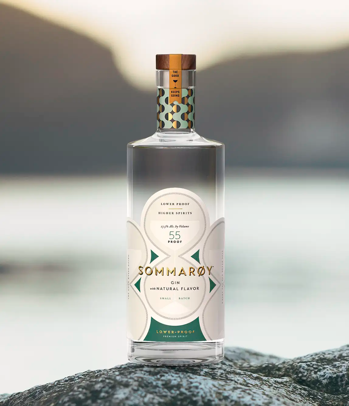A clear bottle of Sommaroy gin with a white label, placed on rocks outdoors. The label indicates it's a lower-proof, natural flavor gin with 55 proof and 27.5% alcohol by volume, designed for small batch production.