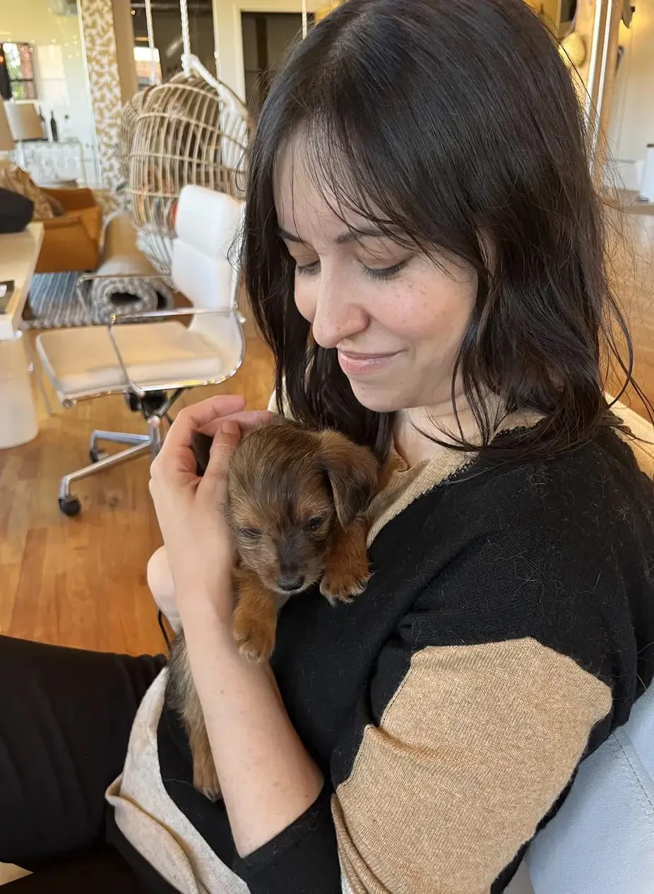 A woman with dark hair holding a small brown puppy close to her chest indoors, smiling gently with her eyes closed.