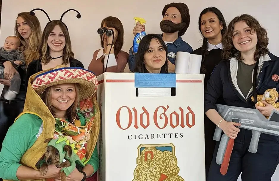 Group of women dressed in costumes for a themed event, with one woman in a bee costume holding a dog, another with a mustache and holding a spray bottle, and others with costumes including a large wrench, surrounded by props and a giant Old Gold ciga