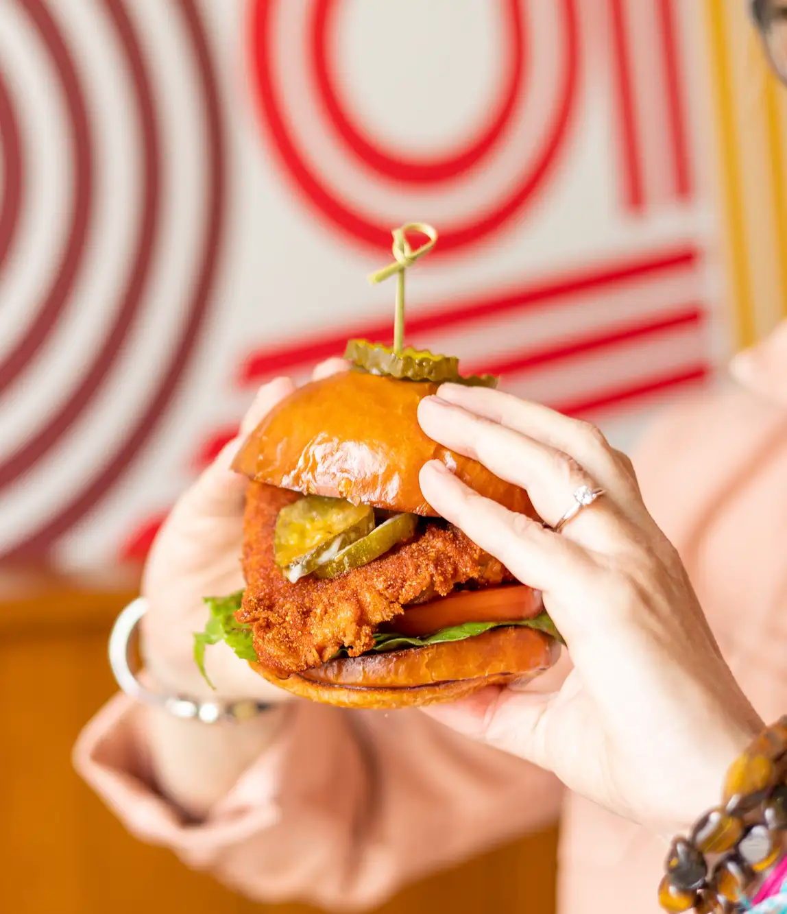 Person holding a fried chicken sandwich with pickles, lettuce, tomato, and a bun, secured with a decorative pick, with a red and white spiral patterned background.