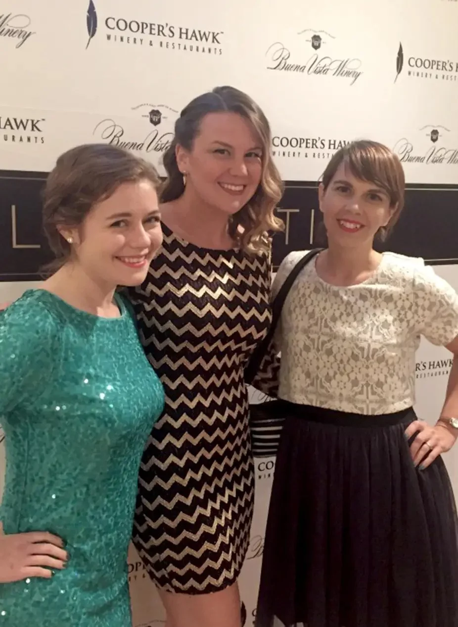 Three women smiling and posing at an event, standing in front of a backdrop with winery names, including Cooper's Hawk and Buona Vita Muray.