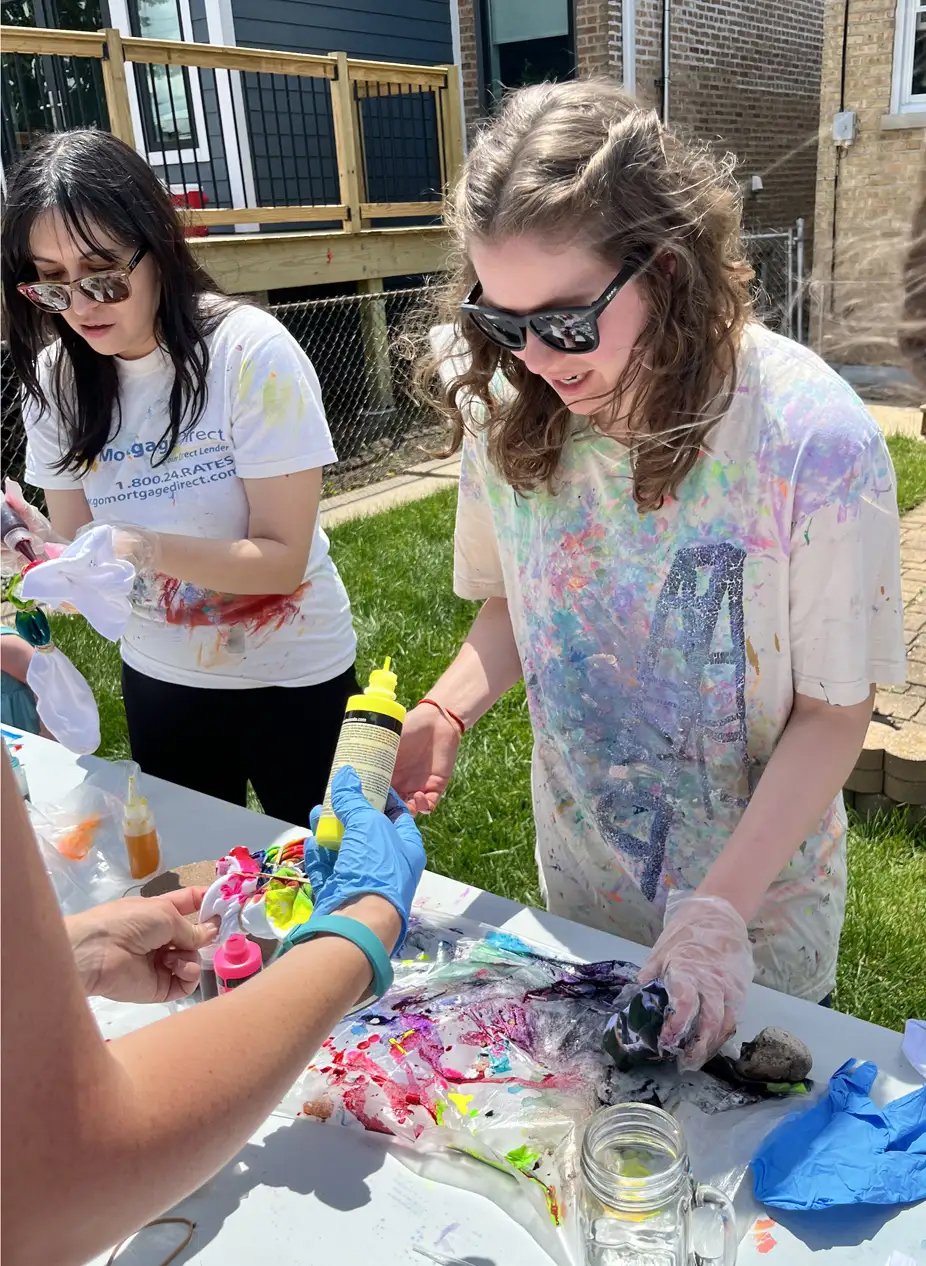 Two young women wearing colorful paint-splattered t-shirts and sunglasses are engaging in a craft activity outdoors at a table covered with art supplies, including paint bottles, gloves, and a rock.