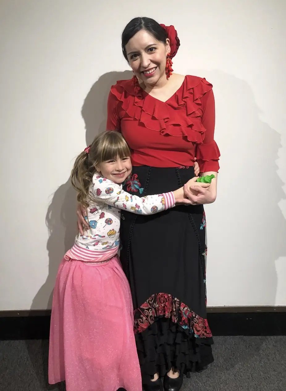 A woman and a young girl hugging and smiling. The woman is dressed in a red blouse with ruffles and black skirt, with dark hair and red accessories. The girl is wearing a colorful sweater with candies and a pink skirt, with blonde hair in pigtails.