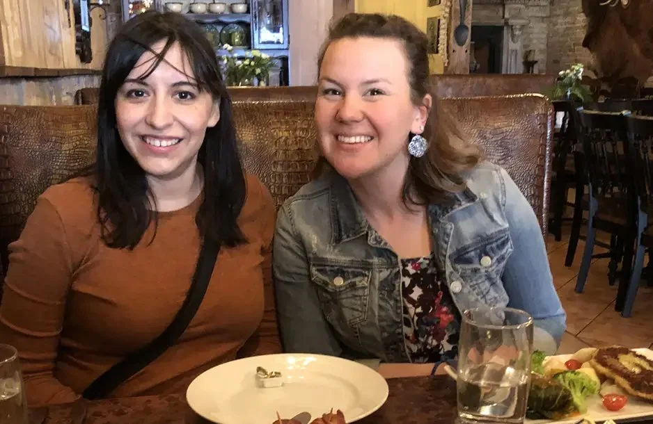Two women smiling and sitting at a restaurant table with food and drinks.