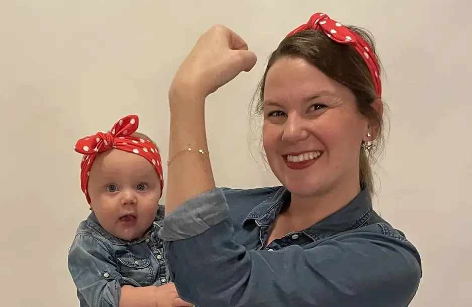 A woman and a baby girl both wearing matching denim outfits and red polka dot headbands. The woman is flexing her arm, and both are smiling.