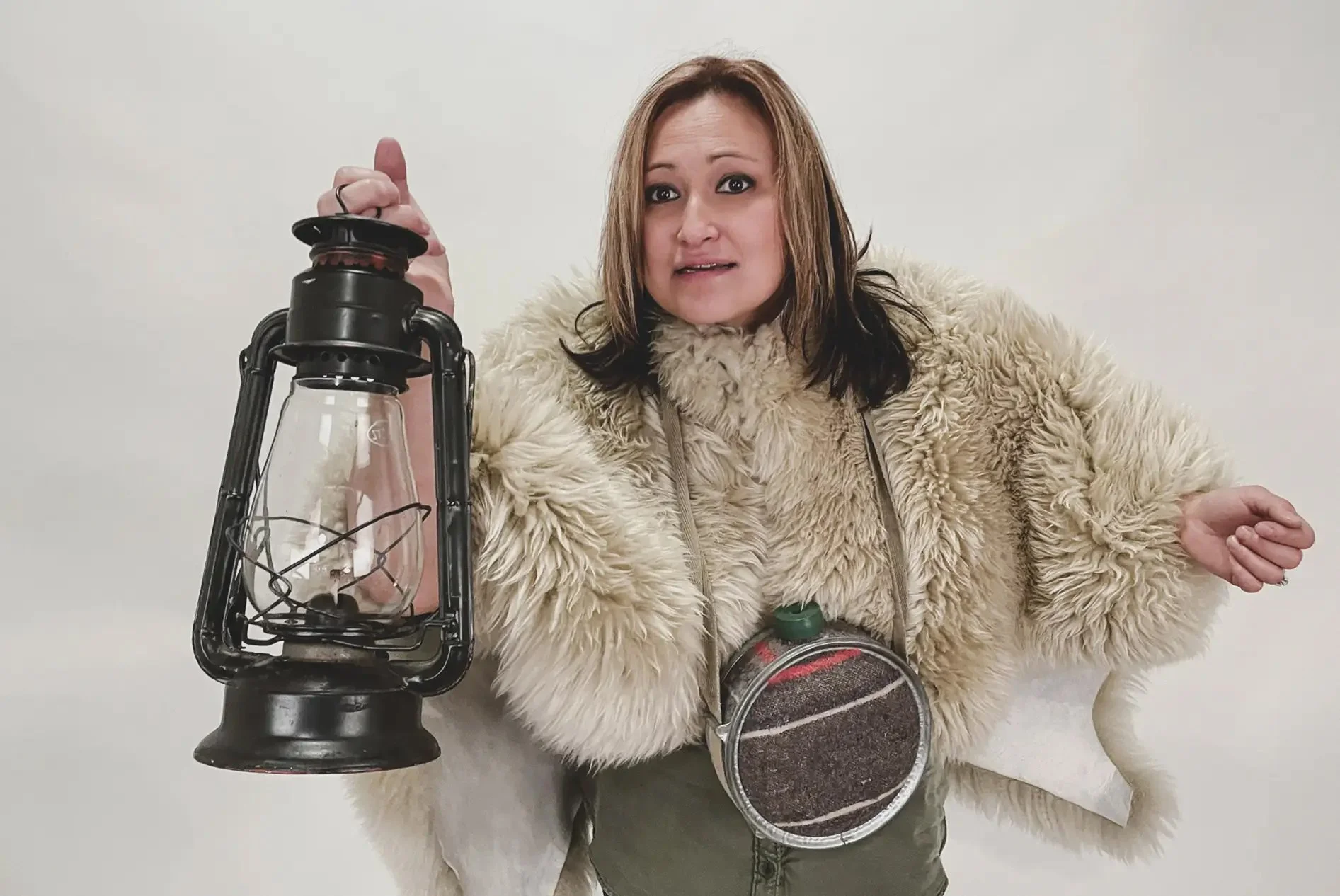 Woman wearing a fluffy coat holding a vintage lantern, with an improvised camera around her neck, standing against a plain white background.