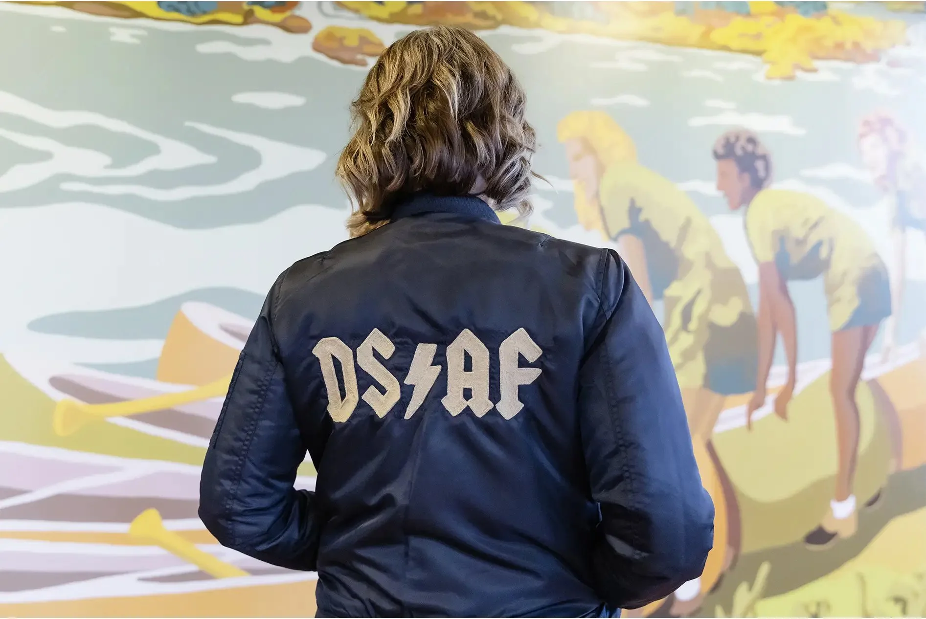 A person with shoulder-length hair wearing a navy blue jacket with 'DSAF' in gold letters on the back, standing in front of a colorful mural depicting children playing near a body of water.