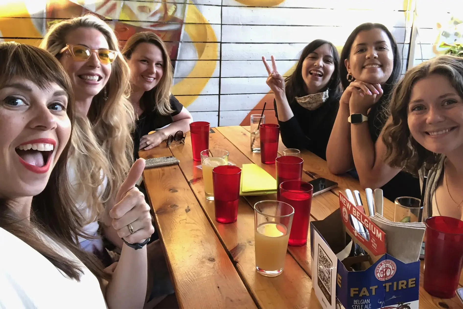Group of six women sitting around a wooden table in a restaurant or cafe, smiling and taking a selfie. The table has drinks, a small box of utensils, and a menu. Bright lighting and colorful wall art are visible in the background.