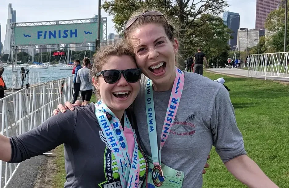 Two women at a finish line, celebrating their race completion, wearing medals and smiling.