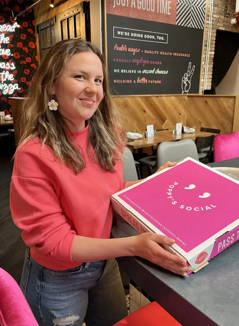 A woman with long wavy hair, wearing a pink sweatshirt and floral earrings, smiling while holding a pink and white box pizza box with a wooden wall and framed chalkboard in the background.