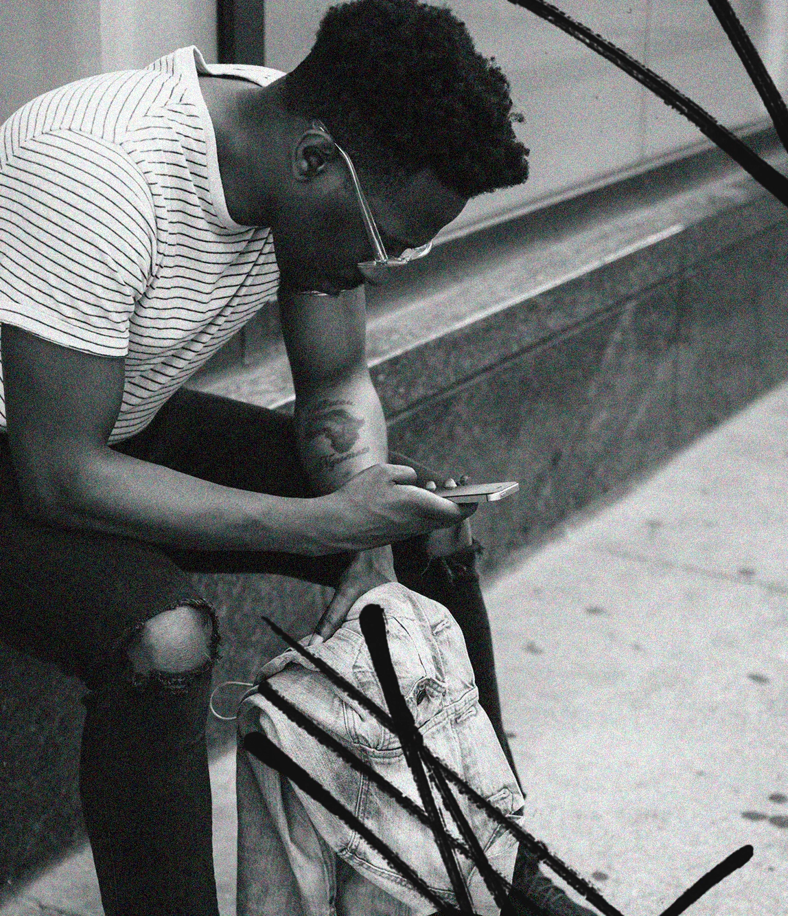 A young man with glasses, wearing a striped t-shirt and ripped jeans, sitting on a bench, looking at his phone, with a backpack on the ground beside him.