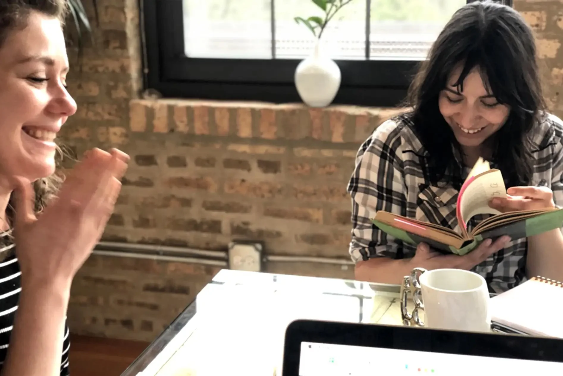 Two women smile and laugh at a table, one holding an open book, in a cozy room with a brick wall, a window, and a potted plant.