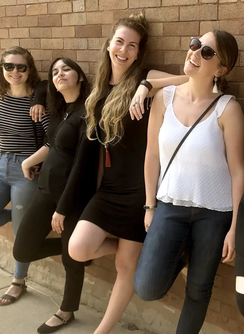 Four women standing against a brick wall, smiling and enjoying the moment, some wearing sunglasses, dressed casually.