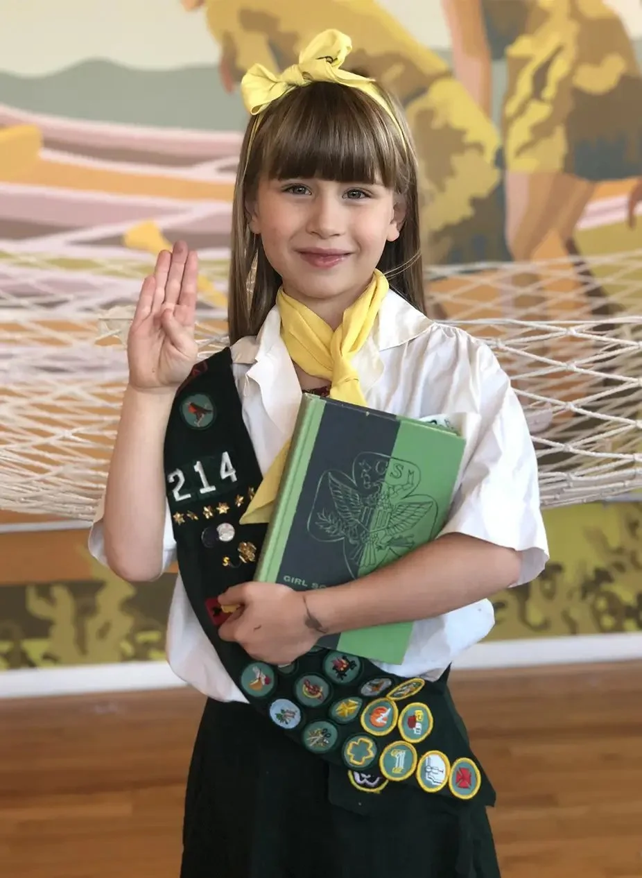 A young girl in Girl Scouts uniform holding a notebook, wearing a yellow neckerchief, and making the Girl Scout sign with her right hand, standing indoors with a colorful mural in the background.