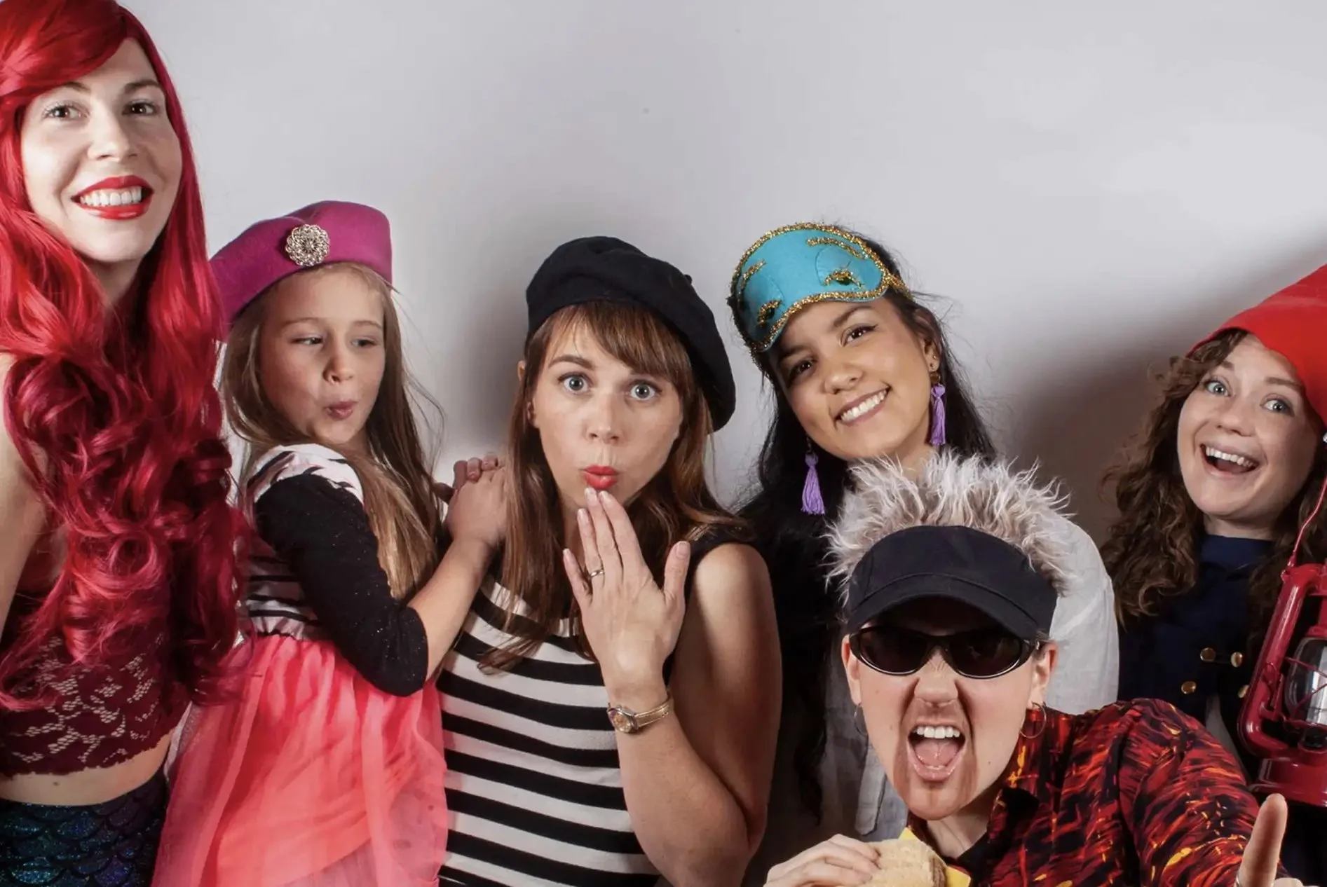 Group of six women and girls dressed in colorful costumes and hats, smiling and posing together against a plain white background.