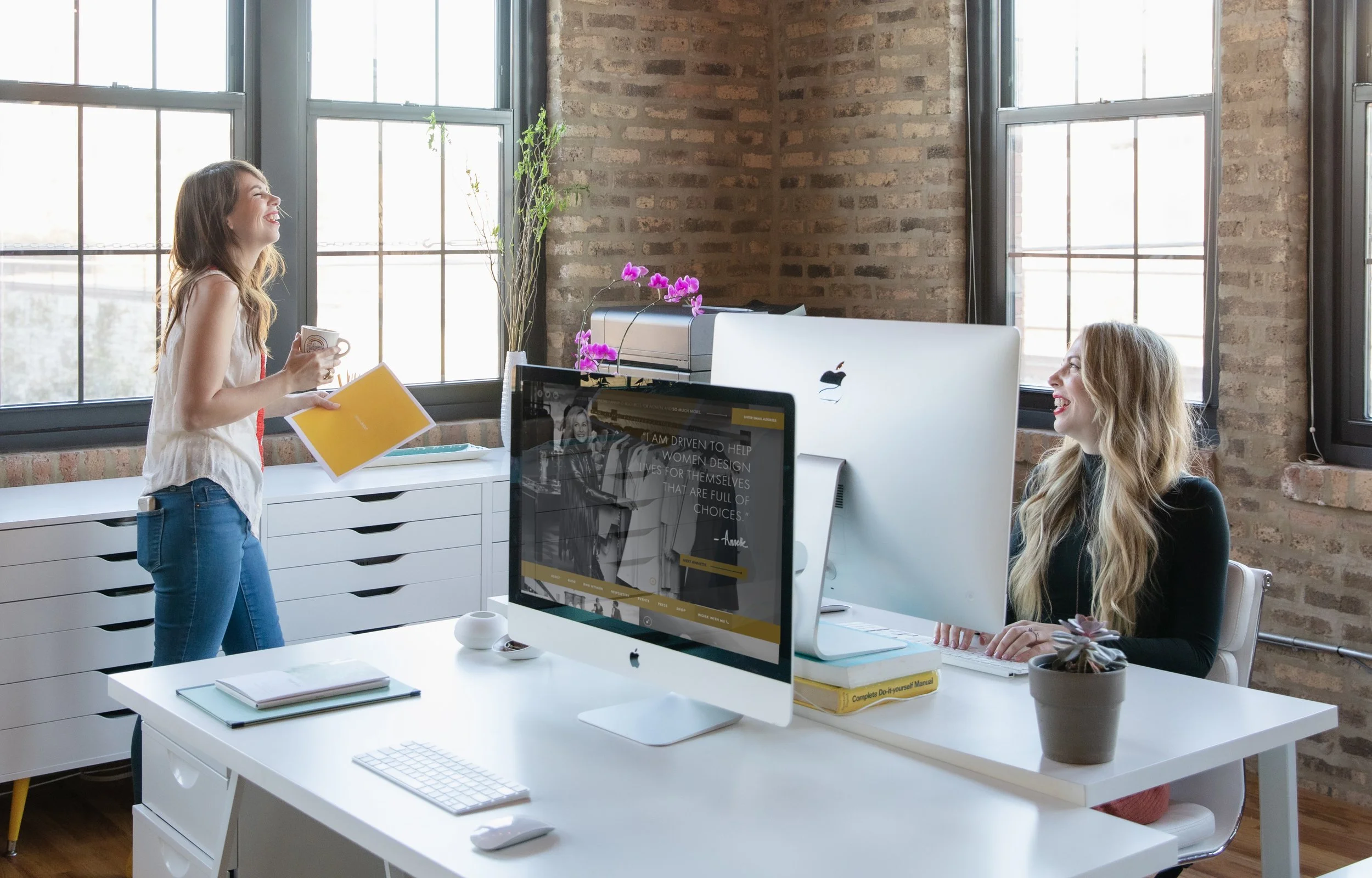 Two women in a modern office with exposed brick walls and large windows, one standing and smiling, holding a mug and a yellow folder, the other sitting at a desk with computer monitors, smiling back, with a plant and a printer nearby.