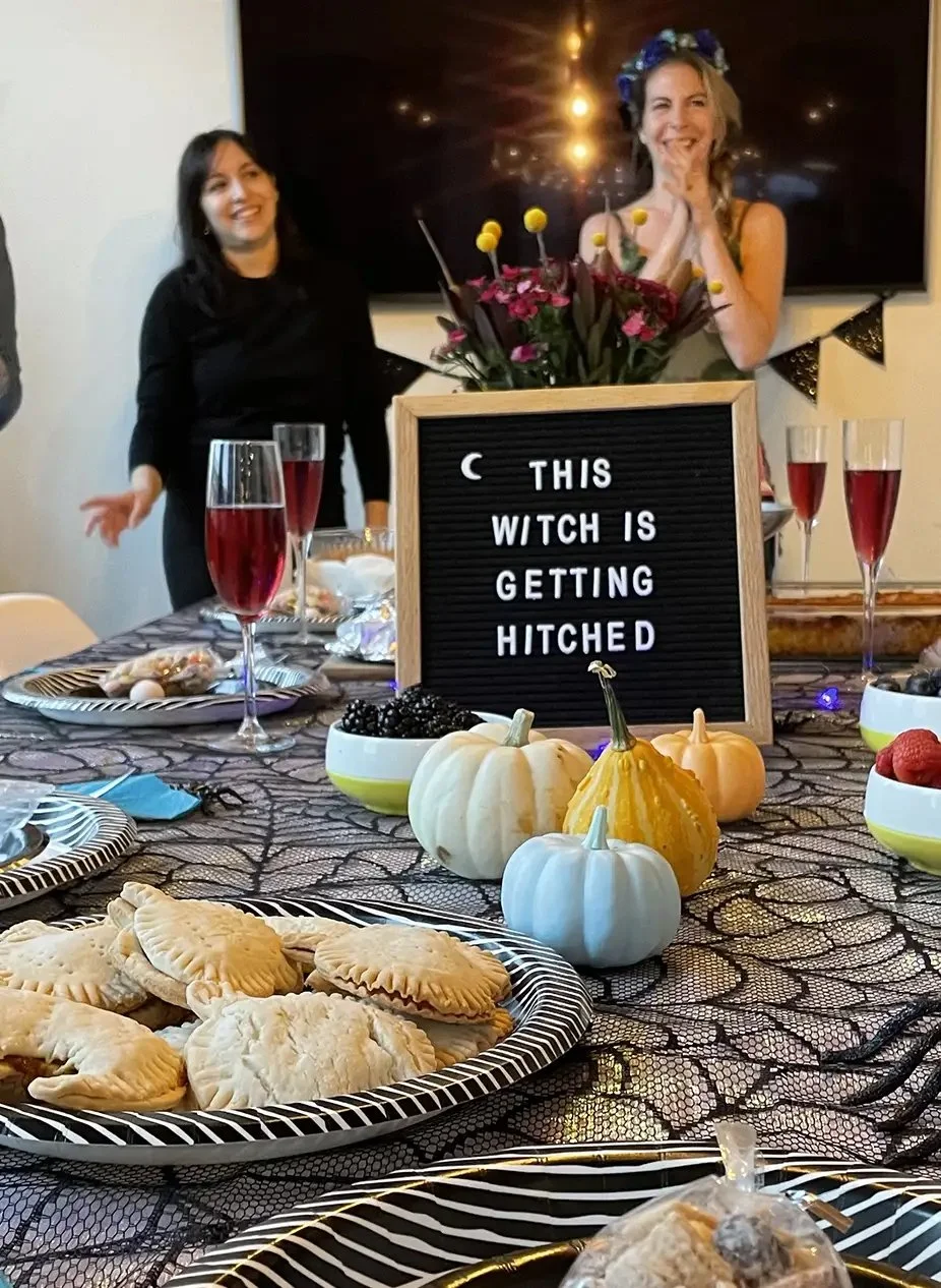 A table decorated for Halloween with a variety of pumpkin decorations, baked goods, and glasses of red beverage. Two women are in the background, one smiling and the other with hands together near her face. A sign on the table reads 'This Witch is Ge