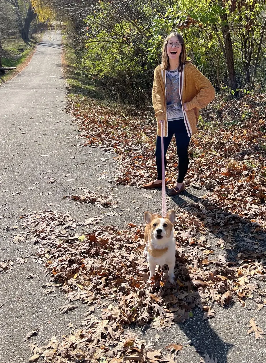 A woman laughing and standing on a leaf-covered path with her corgi dog during fall.