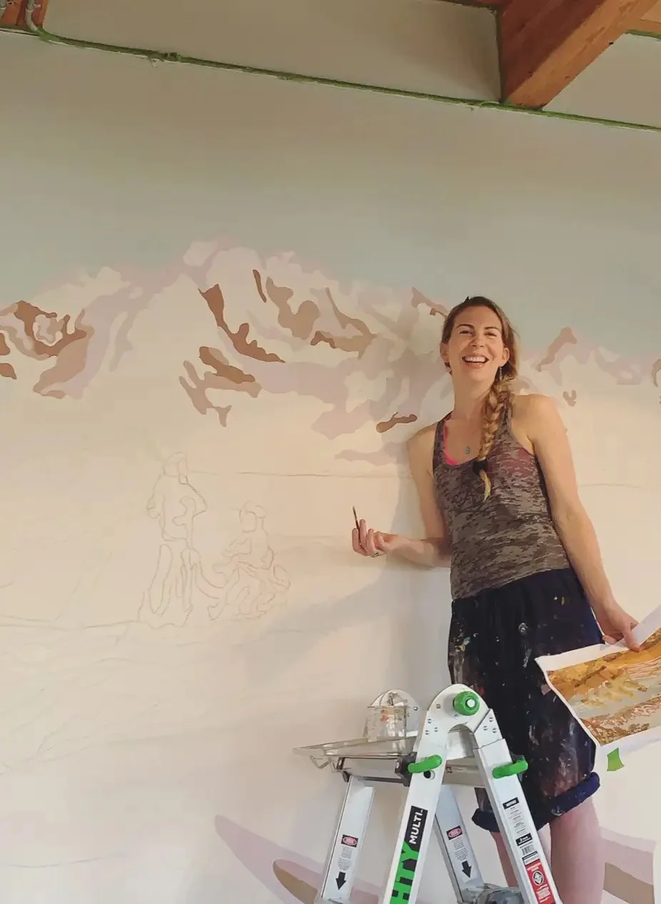 Woman standing on a step ladder, painting a mountain landscape on a wall with a paintbrush, smiling.