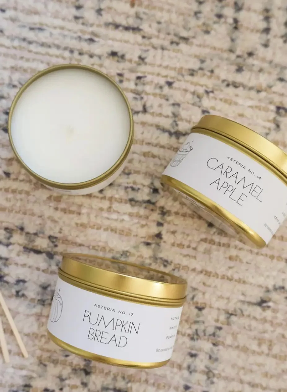 Two candles in white containers with gold lids, one labeled 'Caramel Apple' and the other 'Pumpkin Bread', on a textured beige and brown surface.