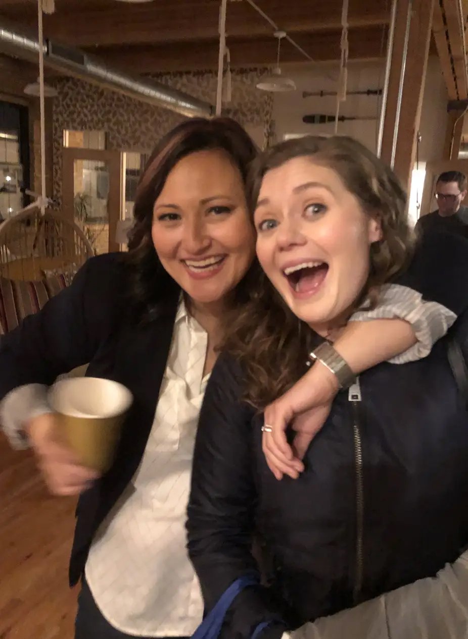 Two smiling women, one holding a yellow cup, in a warmly lit indoor setting with wood beams and hanging decorations.