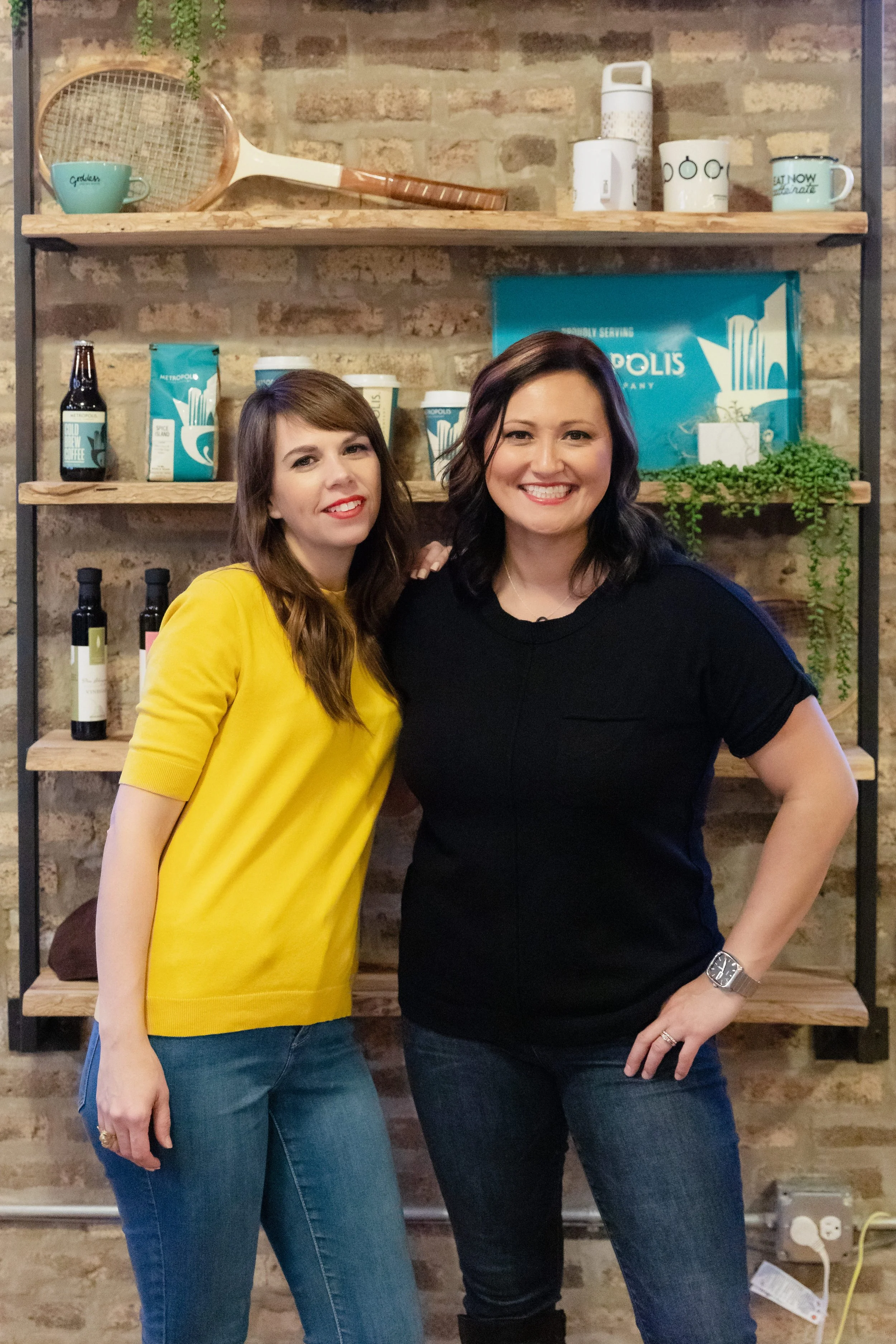 Two women standing close together smiling in front of a brick wall with shelves holding books, bottles, cups, and a tennis racket.